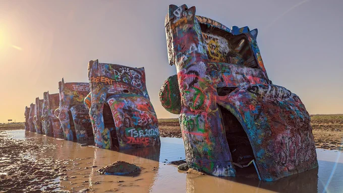 Cadillac Ranch in Amarillo, Texas. Six old Cadillacs with fins stick upward out of a muddy pool of brown water. The cars are thickly coated in spray paint tags of all colors. The sun peeks out from the left.
