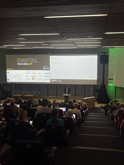 Auditorium full of people looking forward listening to a speaker on stage.