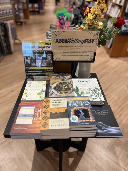 An image of a black square table holding a black-framed header, a poster and 7 books, all piled in a pyramid. The books are, from top left to bottom right: The Hedgerow Handbook by Adele Nozedar, Buds and Blossoms by Liz Knight, Forage by Liz Knight, Bwytai Cymru by Lowri Haf Cooke, Ambassador of Nowhere by Richard Gwyn, Wyneb yn Wyneb by Sioned Wyn Roberts and Skirrid Hill by Owen Sheers. The header and poster is promoting the Abergavenny Writing Festival.