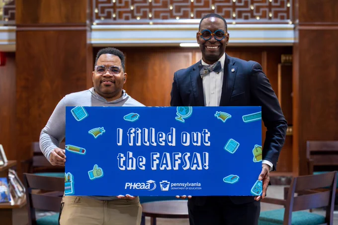 The Secretary of Education and a student hold up a blue sign that reads, "I filled out the FAFSA" with the PHEAA and PDE logos on it. 