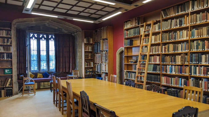 Library of the Week. Magdalen College. United Kingdom. Interior shot of a room with a long wooden table, crammed bookshelves, and gothic windows.