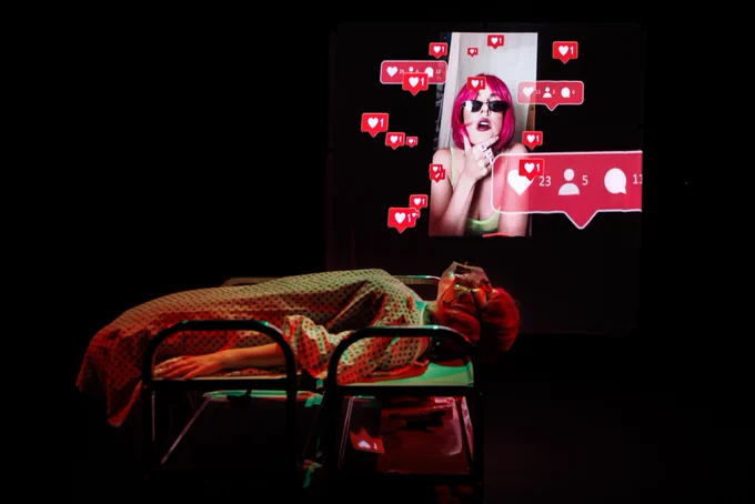 a woman wearing a hospital gown and oxygen mask lies down across medical looking catering trolleys. In the dark behind her is a projected image of her in a pink wig, on instagram live, with multiple love heart and like symbols around her.