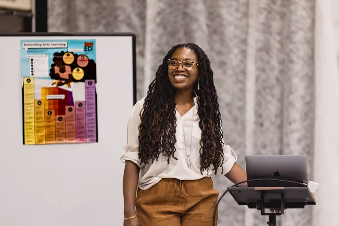 Alecia Dawn stands smiling between a laptop podium and a white board featuring Arts Ed Collaborative's theory of change poster (that she helped illustrate!).