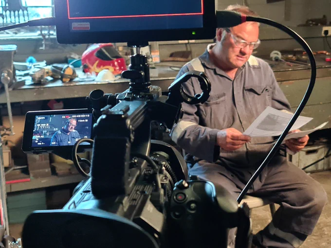 Photo of a person wearing a grey boiler suit sitting in a chair in a workshop and reading some papers. There is a camera on a tripod in front of him and filming him.