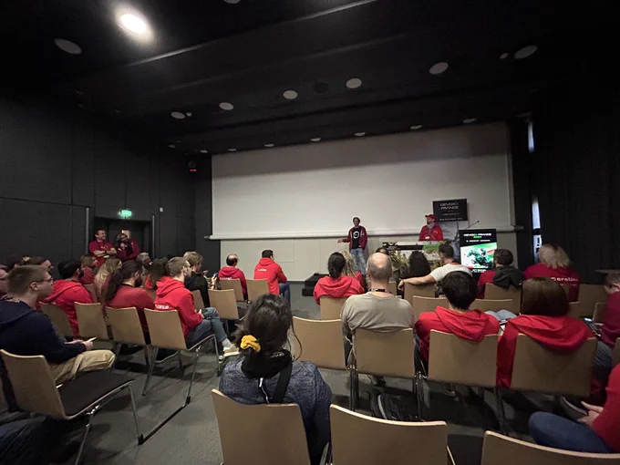 Ensemble de 40 personnes assis dans une salle de conférence avec un speaker sur la scène. Presque tout le monde porte un polo rouge.