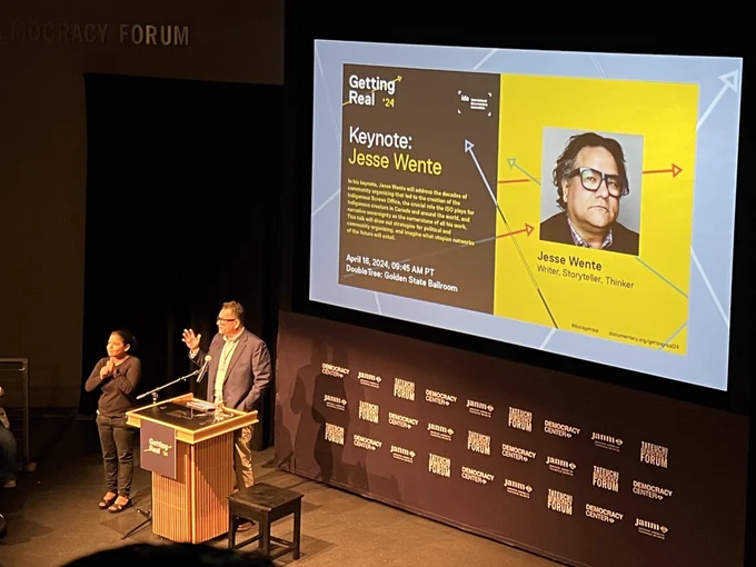 Photo of Jesse went middle aged indigenous man with glasses in a suit behind a podium with an interpreter wearing black clothing. They are standing in front of a screen with text that reads Jesse Wente and getting real 24 logo.