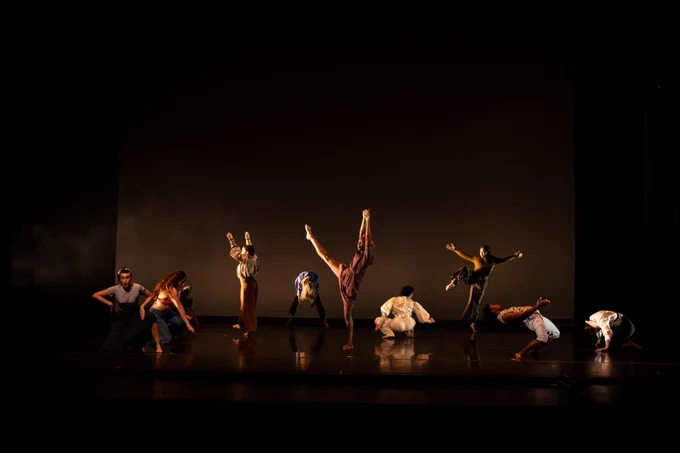 The photo shows nine dancers on a dimly lit stage. On the far left, one crouches in gray attire, while the next, facing right, also crouches. Further back, a dancer raises both hands, while another bends forward with dangling hair. Downstage, one extends a leg and arms. A dancer in all white faces away, another stands on toe with extended arms. Finally, two in white; one leans back with raised arms, the other crouches in a huddle. Photo by Miya Tanner. 