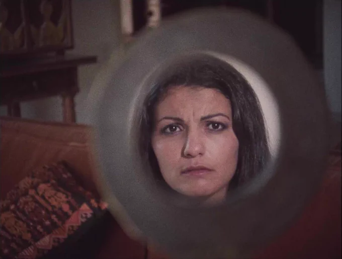 A still from the film Leila & the Wolves, in which a brown woman of Arab origin looks at her own reflection in a circle-shaped mirror. Her hair is dark and her face expresses discomfort. What surrounds the mirror is a domestic-setting with partially visible furniture.