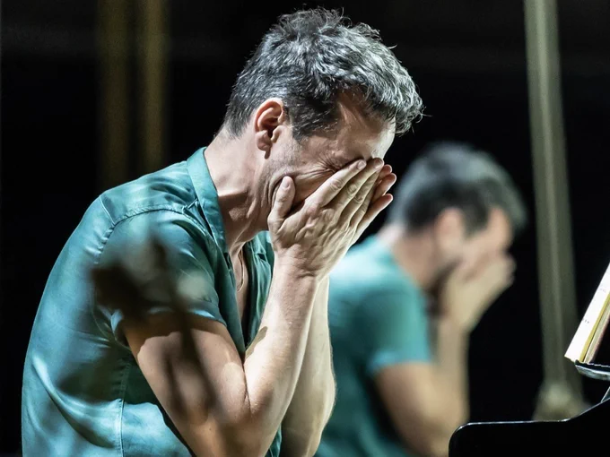 A man in a blue shirt holds his face in his hands as if crying. Sat at a piano.
