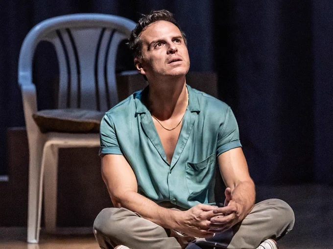 A man in a blue shirt and chinos sits cross legged on the floor and looks contemplative. A white plastic chair behind him.