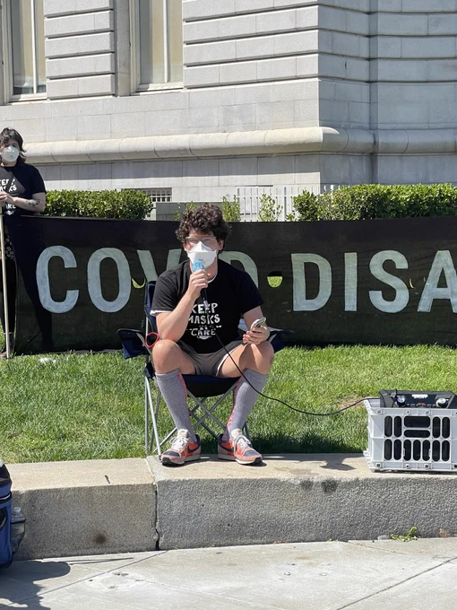 White man sitting in a folding chair and holding a microphone. Behind him is a banner that says COVID DISABLES