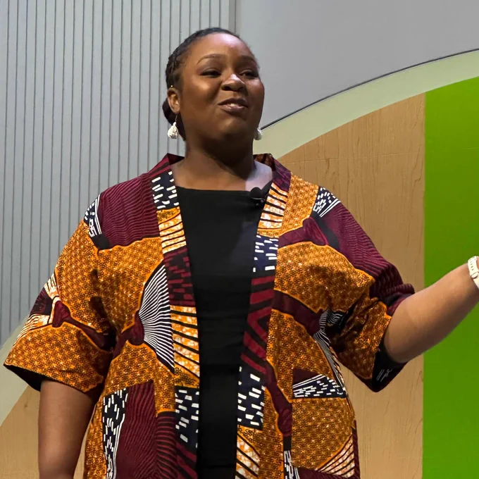 Ajuna smiling while talking on a stage in an African print kimono style jacket and black pants.