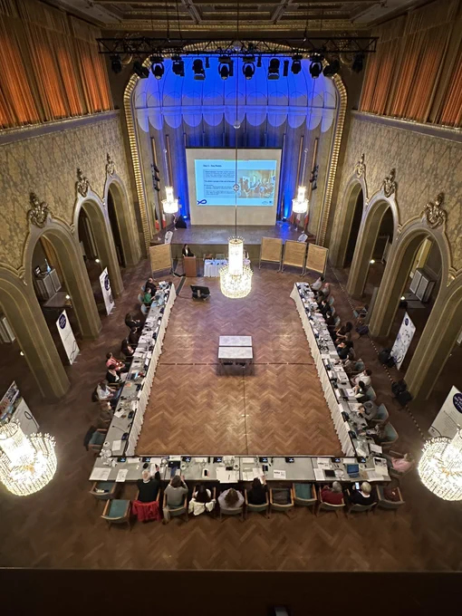 A group of people sitting around a u-shaped table looking at a presentation in an ornate room