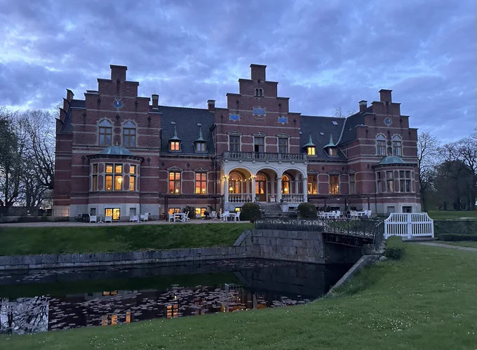 Photograph of a 19th century manor house, at dusk: a beautifully symmetrical red brick building in a mix of styles, with a lily-pad strewn moat cutting through the verdant grass.