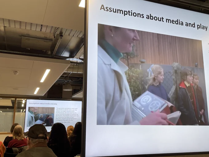 'Assumptions about media and play'. A side by side view of the screen with the video of children at a playground, and the audience watching another screen.