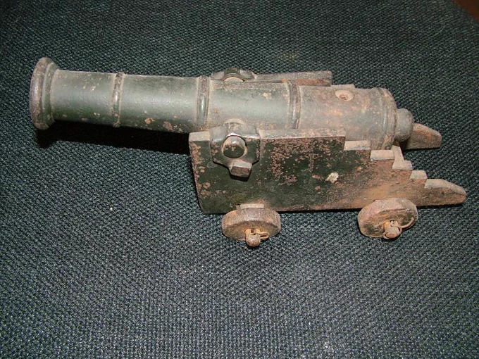 A miniature cannon. It has a long cylindrical barrel pointing to the left. The end of the barrel is flared. In the middle of the barrel there is two small iron cylinders sticking out either sides. This is attached to a moveable base with two wheels either side. The entire object is dark grey/black, with patches of dark coral pink/orange where the colour has faded.