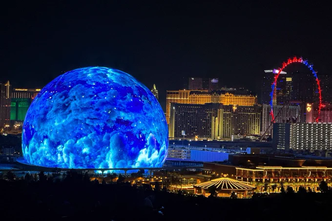 Photo of the MSG Sphere in Las Vegas at night. The Sphere is shown in the left portion of the photo. The exterior is showing a pattern that looks like clouds on a deep blue background.  The High Roller Ferris wheel is illuminated in red and blue on the right hand side of the photo. In the center background of the photo, Bally’s and Paris hotels can be seen.