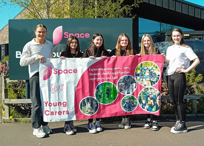 Youth worker Robbie, wearing a white long-sleeve shirt and black jeans, stands outside Space with a group of five young people, all holding a pink-and-white banner promoting Young Carers.