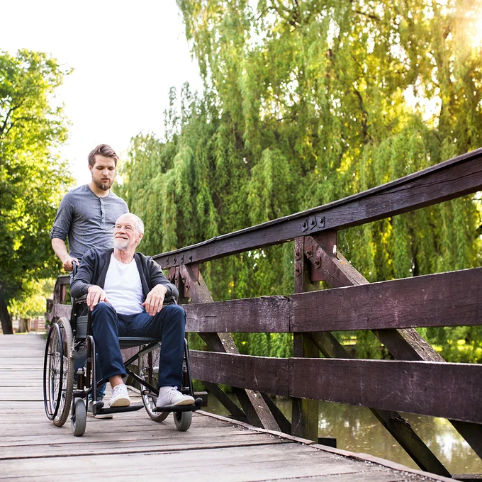 Älterer Mann im Rollstuhl wird von jüngerem Angehörigen über eine Holzbrücke im Park geschoben