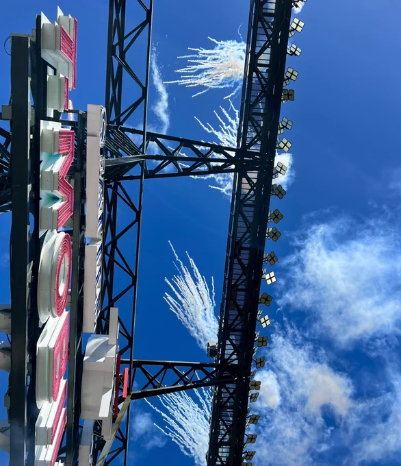 Photo directly overhead from left field bleachers, with the stadium lights bisecting a deep blue sky. Part of the “T-Mobile” branding is visible from beneath. The white smoky tendrils of recently-exploded fireworks are visible in the sky, directly overhead. Wispier smoke blows off to the right hand side of the image.
