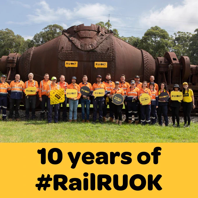 Rail workers pose with R U OK?staff holding signs and flags. Text reads: 'Ten years of Rail R U OK?'