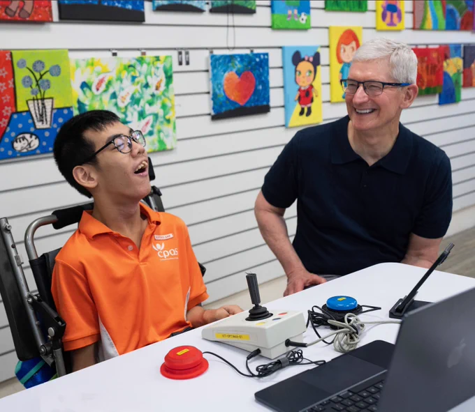 Tim and a student from the Cerebral Palsy Alliance School Singapore sit next to each other at a desk in a classroom, with MacBook open. Student artwork is behind them on the wall.