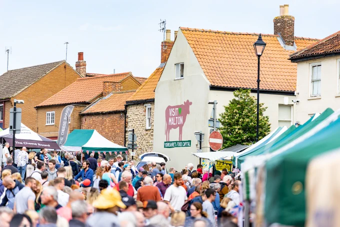 Pictured: Malton Food Lovers Festival (Spring) in full swing taking place around the historic Market Place. Visitors browsing artisan food stalls with views of the Visit Malton cow mural on the wall at Newgate. 