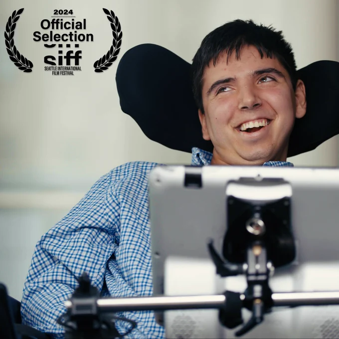 Samuel smiling in his wheelchair with his communication device in front of him. He is wearing a blue checkered button down shirt and is looking up to the left. In the upper left corner above him is a black laurel that reads “2024 Official Selection SIFF Seattle International Film Festival”