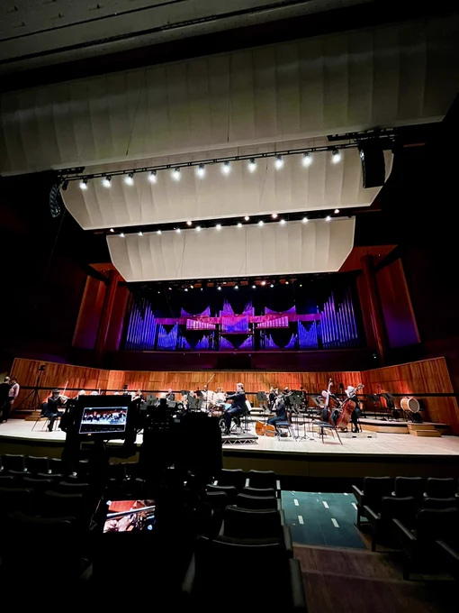 The interior of Royal Festival Hall in London as an orchestra assembles for rehearsal on the dramatically lit stage. In the foreground a television camera waits for the rehearsal to begin.