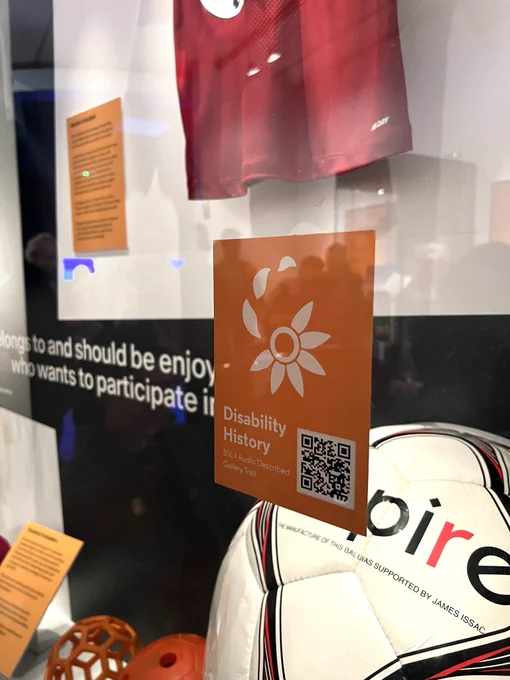 A Disability History trail logo and QR code on an orange sticker. The sticker is on a glass display case. There are sports balls displayed behind the case.