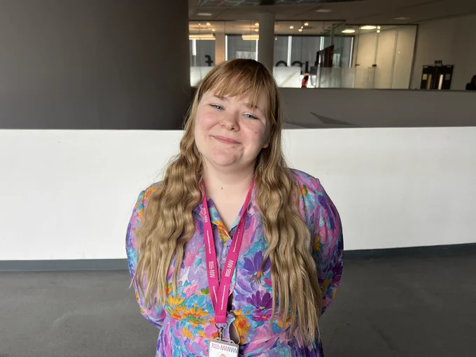 A white woman with long dark blonde hair. She is wearing a colourful floral dress.