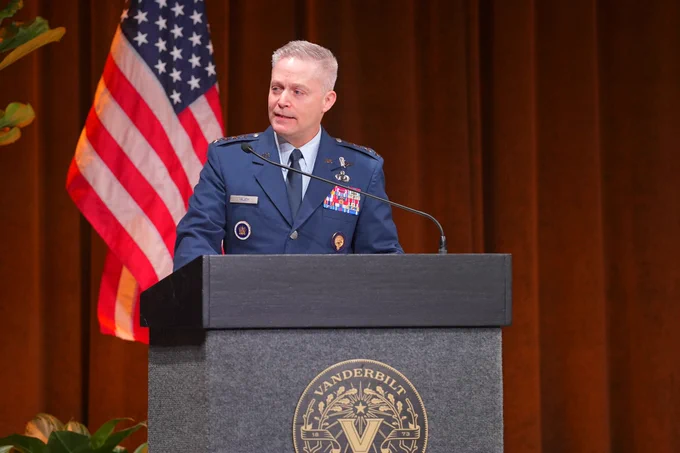 A man in military uniform stands behind a podium, with the American flag hanging behind him.