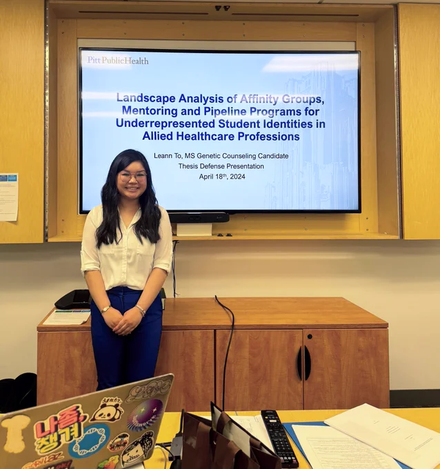 Leann standing in front of a TV monitor showing powerpoint title slide "Landscape Analysis of Affinity Groups, Mentoring and Pipeline Programs for Underrepresented Student Identities in Allied Healthcare Professions"