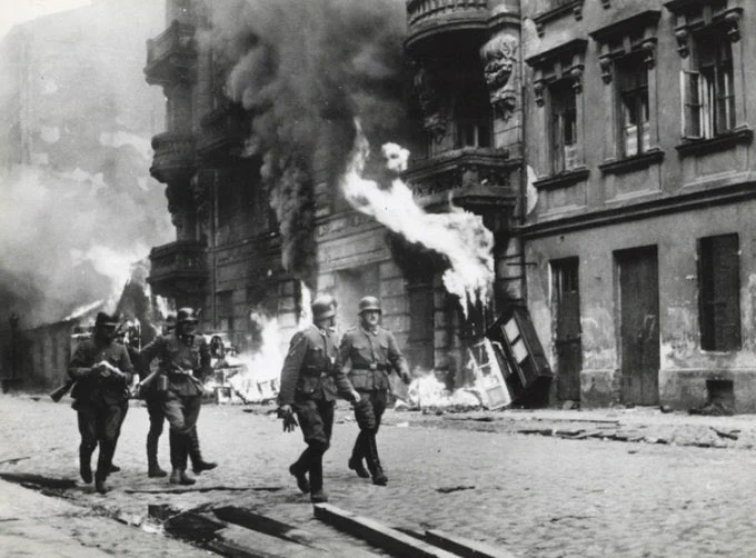 A photo from the Warsaw Ghetto Uprising. A group of German soldiers walking along a street. Behind them a building on fire.