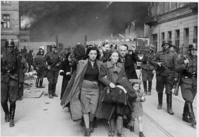 Group of women and children escorted by German soldiers during the Warsaw Uprising. Smoke of the burning city visible in the background.