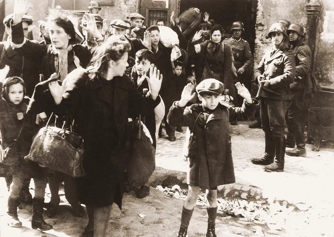 Group of civilians at the Warsaw ghetto uprising. Some people raise their hands up. German soldiers are visible in the background with their guns.