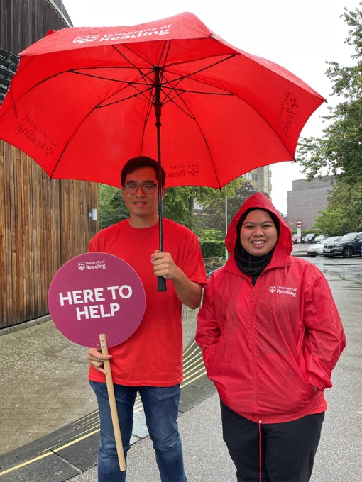 Two University of Reading students wearing branded red clothing stand under an umbrella on a wet and rainy open day.