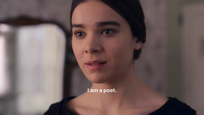 A close-up of Hailee Steinfeld as Emily Dickinson in Dickinson. She has her hair tied in a bun at the back of her head and wears a black top. She stands in a bedroom with floral patterned wallpaper and a mirror on the wall. She has an insistent look on her face. Text on image indicates she is saying, "I am a poet."