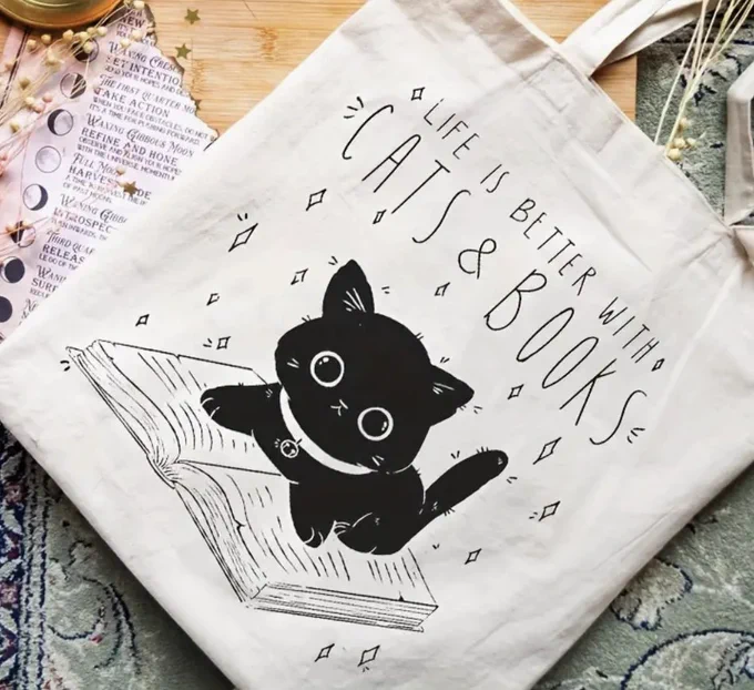  A white tote bag with the image of a black cat reading and the text, "Life is better with Cats & Books".