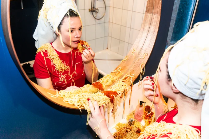 A white woman in a red t-shirt and a head towel looks at her reflection in the mirror and applies red lipstick. Her body and the mirror are also covered in spaghetti.