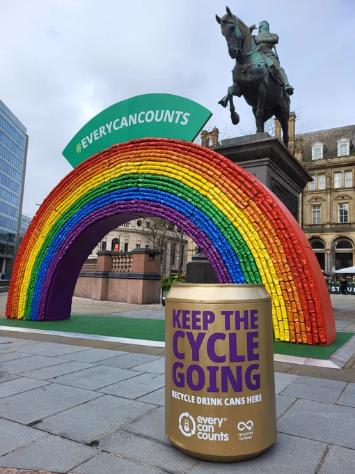 A rainbow arch made of 2,500 drink cans with a statue of the Black Prince behind it all located within City Square, Leeds. In the foreground is a gold can shaped recycling bin.