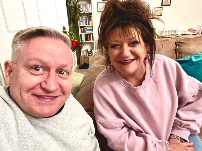 Close up selfie of Simon in a grey jumper and short hair next to Jane with a light purple sweat top and loose dark hair. They are in their living room, a thin bookshelf in the distance. 