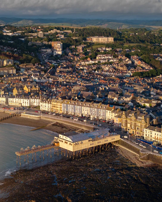 An aerial view of Aberystwyth on the west wales coast with the royal pier in the foreground. The low sun is lighting up the fronts of all the houses. The National library of Wales is visible on the hill in the background with the Cambrian Mountains in the distance.