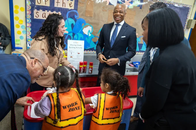 mayor smiles while touring classroom with young girls below him doing STEM demonstration and city officials and teachers standing around them