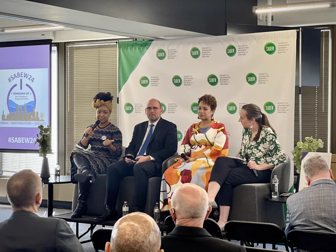 Four people sit next to each other on a small stage with the woman on the far left speaks into a microphone. A screen projection in the background reads, “#SABEW24.”