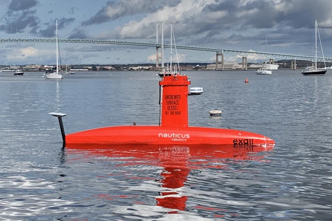 A small vessel shaped like a submarine floats in calm ocean waters. In the background is a large suspension bridge and some sailboats.