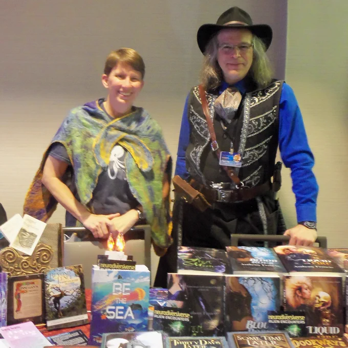 Clara Ward, in a felted sea cloak, stands smiling next to Anthony Francis, dressed finely with a decorated vest. Many books are on the table, many with promotional bookmarks for neurodiversive.