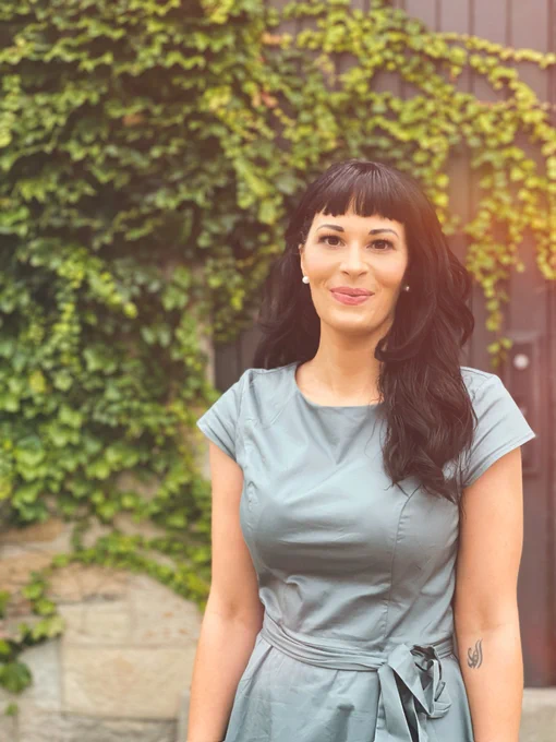 Portrait of a Christina, a white woman standing in front of a wall of green foliage. She’s smiling and has long black hair with fringe bangs, and a grey dress.