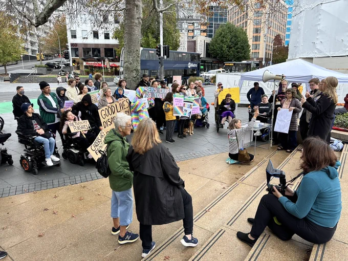 a shot of aotea square showing a few dozen people gathered in front of a stage with a megaphone. The group of people include a range of ages, ethnicities and wheelchair users. People are holding signs such as “return my rights”, “no cuts”, “carers are frontline workers”.