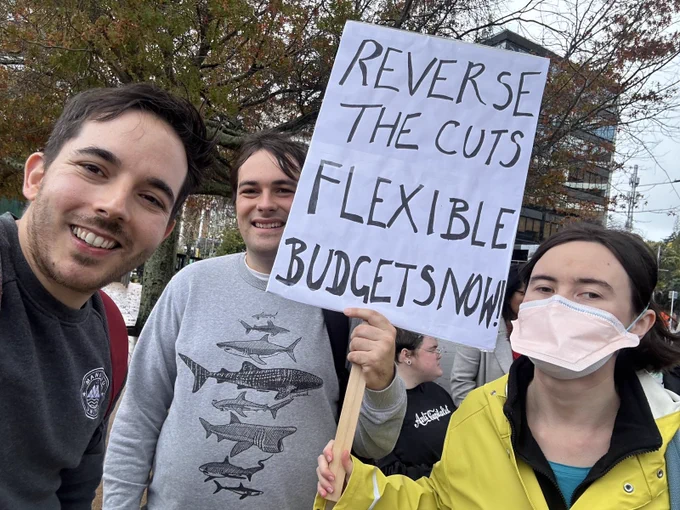 selfie taken by Ricardo showing Ricardo, Jonty, and Áine smiling at Aotea Square. Jonty and Áine are holding a sign that says “Reverse the cuts. Flexible budgets now”.